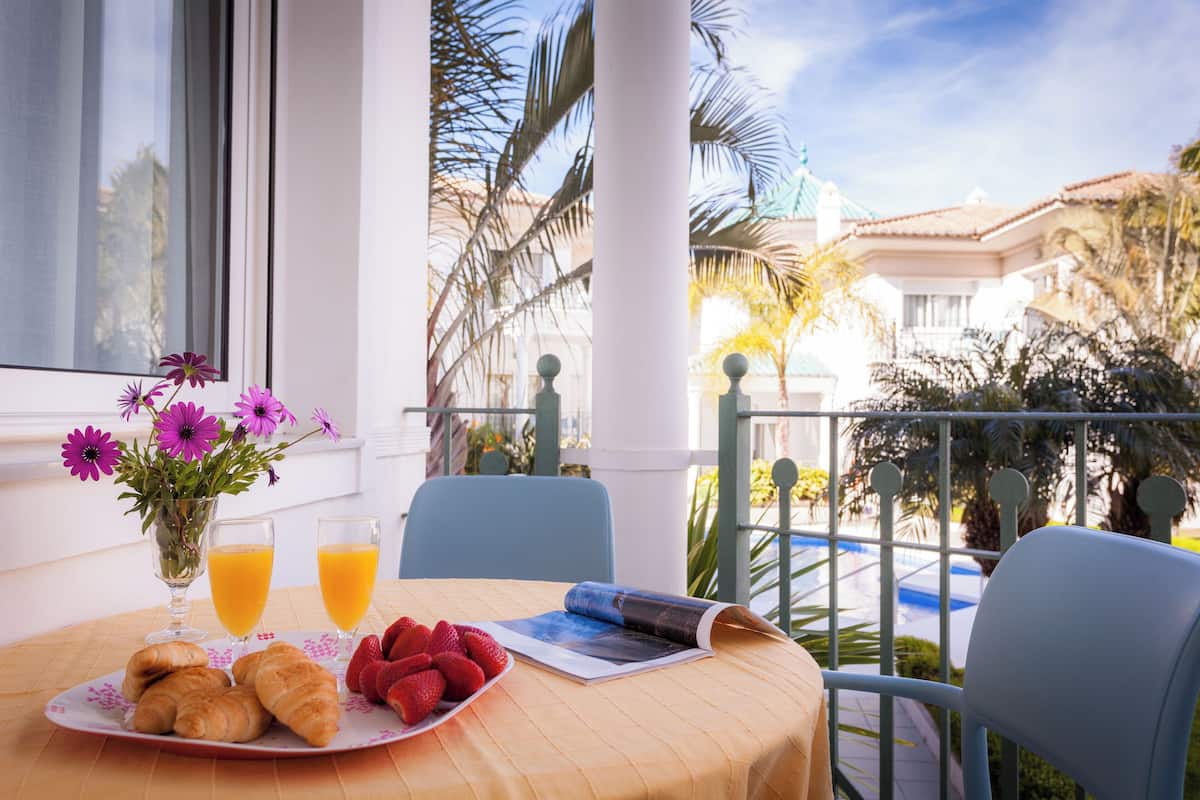 Una mesa con cruasanes, fresas y vasos de jugo, un florero con flores y una revista en un balcón con vista a palmeras y una piscina.