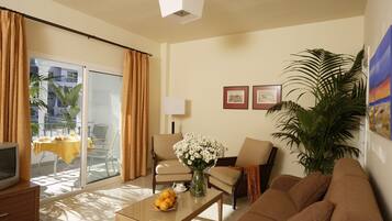 Habitación de hotel con balcón, un sofá, una mesa de centro con fruta, televisor y lámpara.