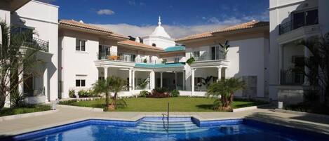A large, white building with a central dome and multiple balconies, surrounded by a swimming pool.