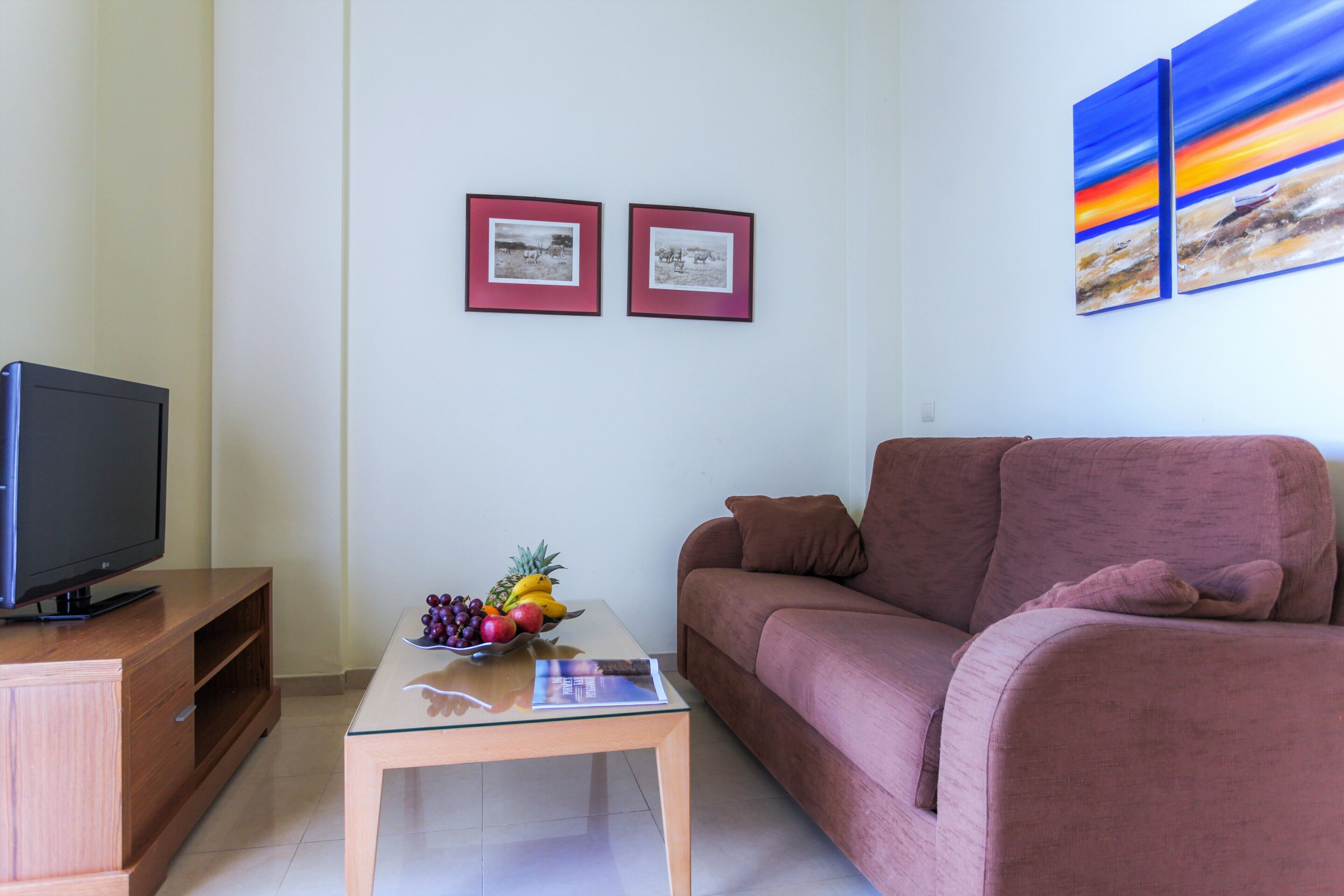 A living room with a television on a wooden cabinet, a glass coffee table with fruit and a magazine, a brown sofa, and two framed pictures on the wall.