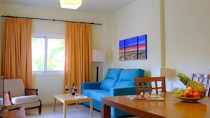 Ein Wohnzimmer mit einem blauen Sofa, einem Holztisch, einem Esstisch mit Obstschale und einem Gemälde an der Wand.