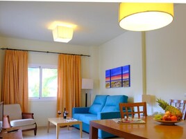 A living room with a blue sofa, a wooden coffee table, a dining table with a fruit bowl, and a painting on the wall.