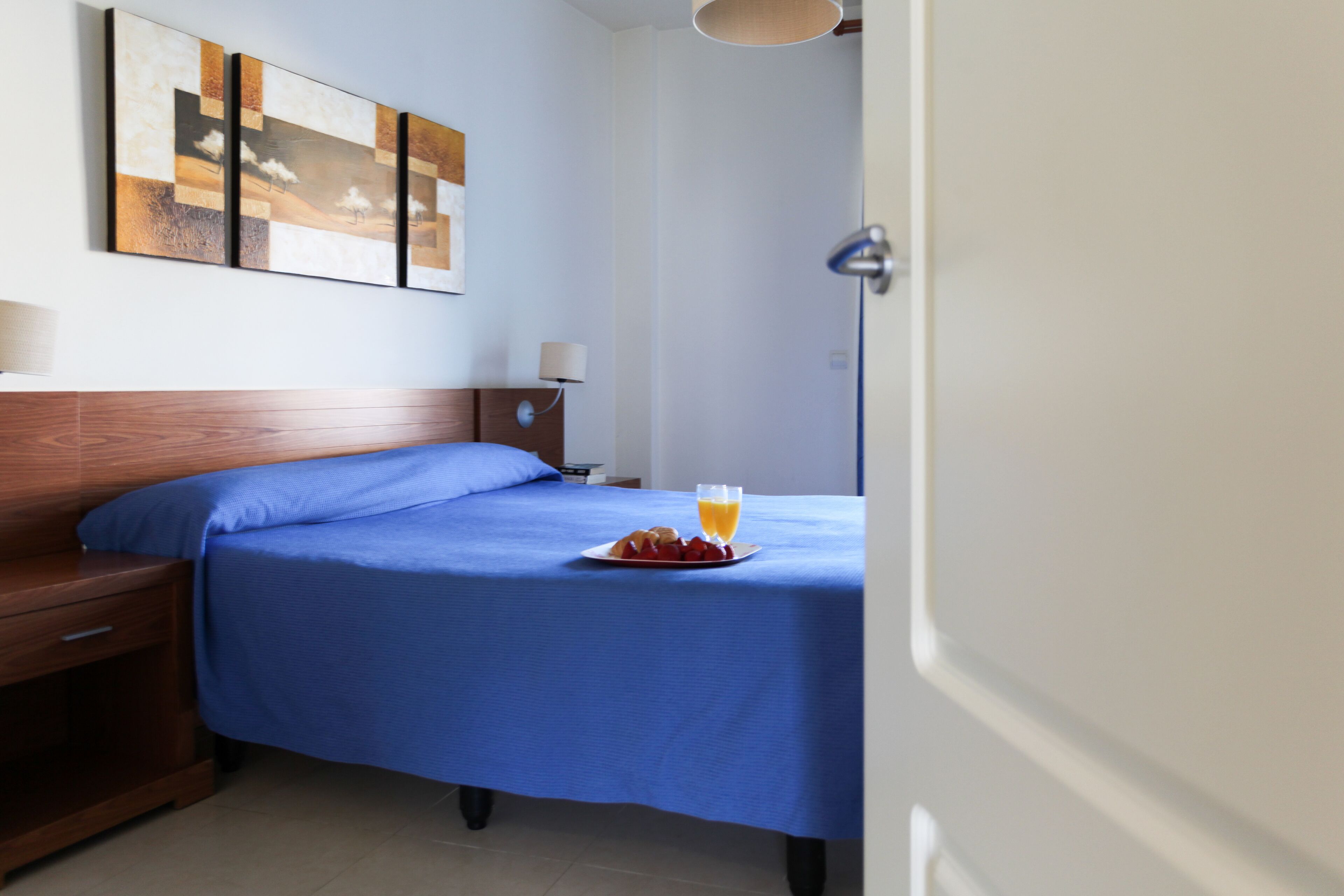 A neatly made bed with a blue bedspread, a bedside table with a lamp, and a plate of fruit and juice on the bed.