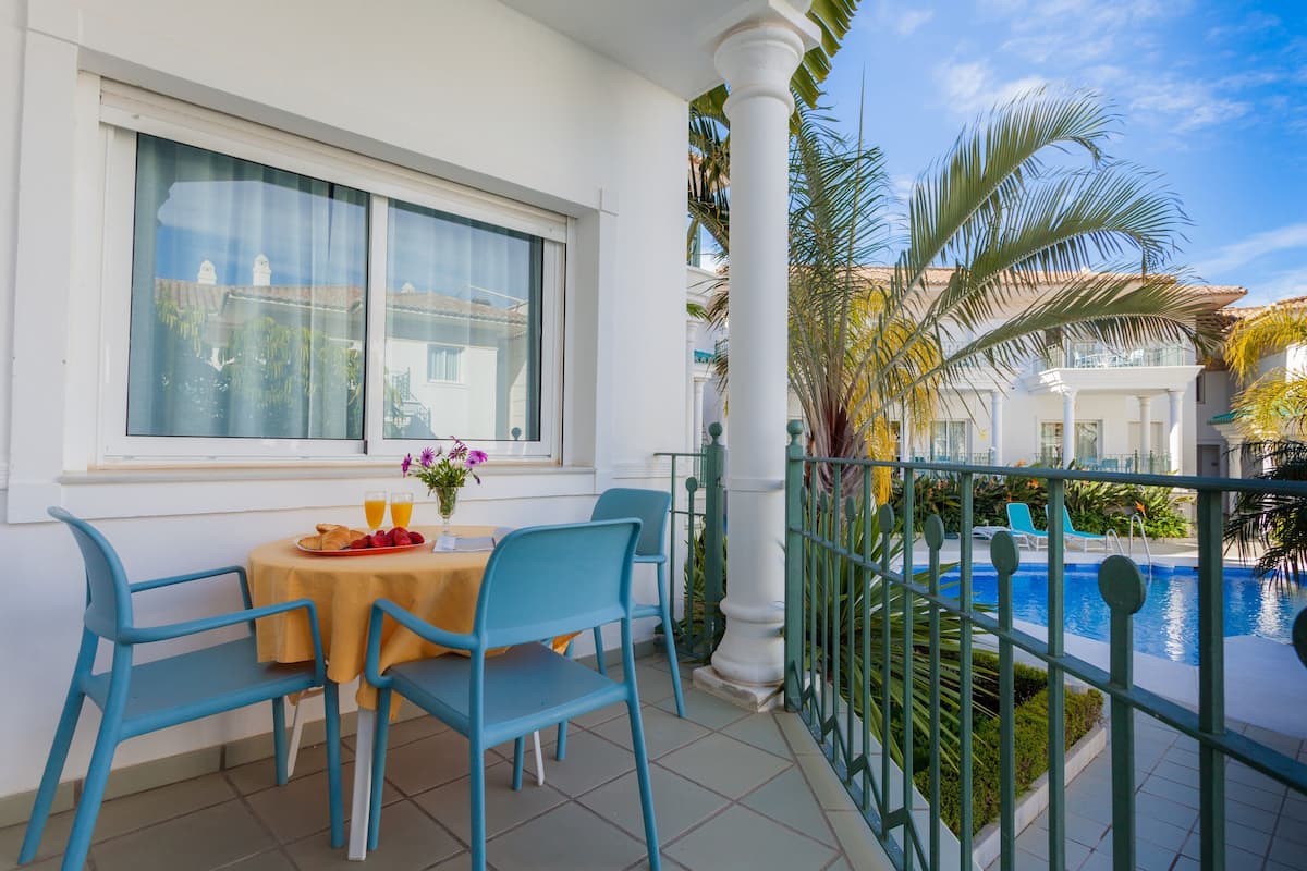 Un balcón con una mesa dispuesta para el desayuno, una piscina y vistas a un edificio de fachada blanca.