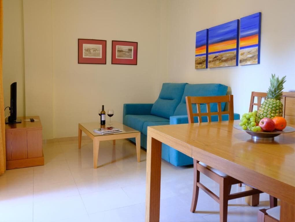 Uma sala de estar com um sofá azul, uma mesa de jantar de madeira e uma tigela de frutas. Há quadros nas paredes e uma máquina de café em uma mesa lateral.