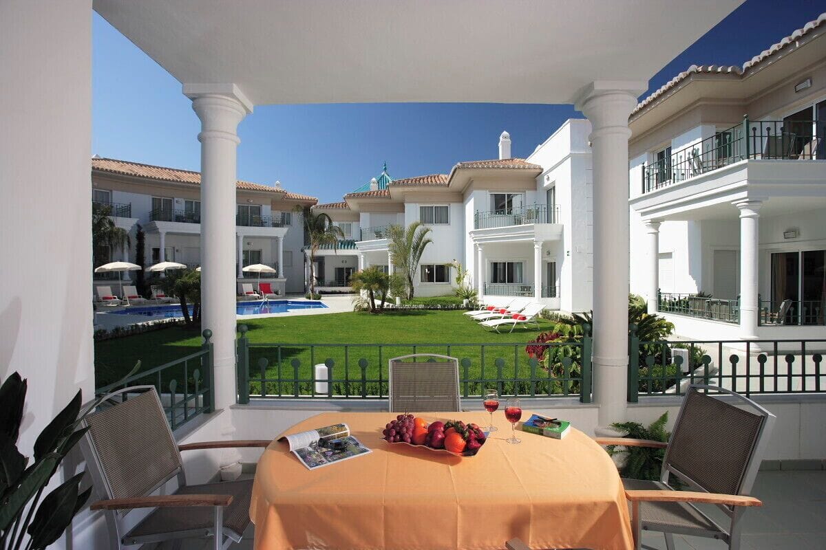 Un balcón con una mesa para dos, con vistas a una piscina y un césped cuidado.