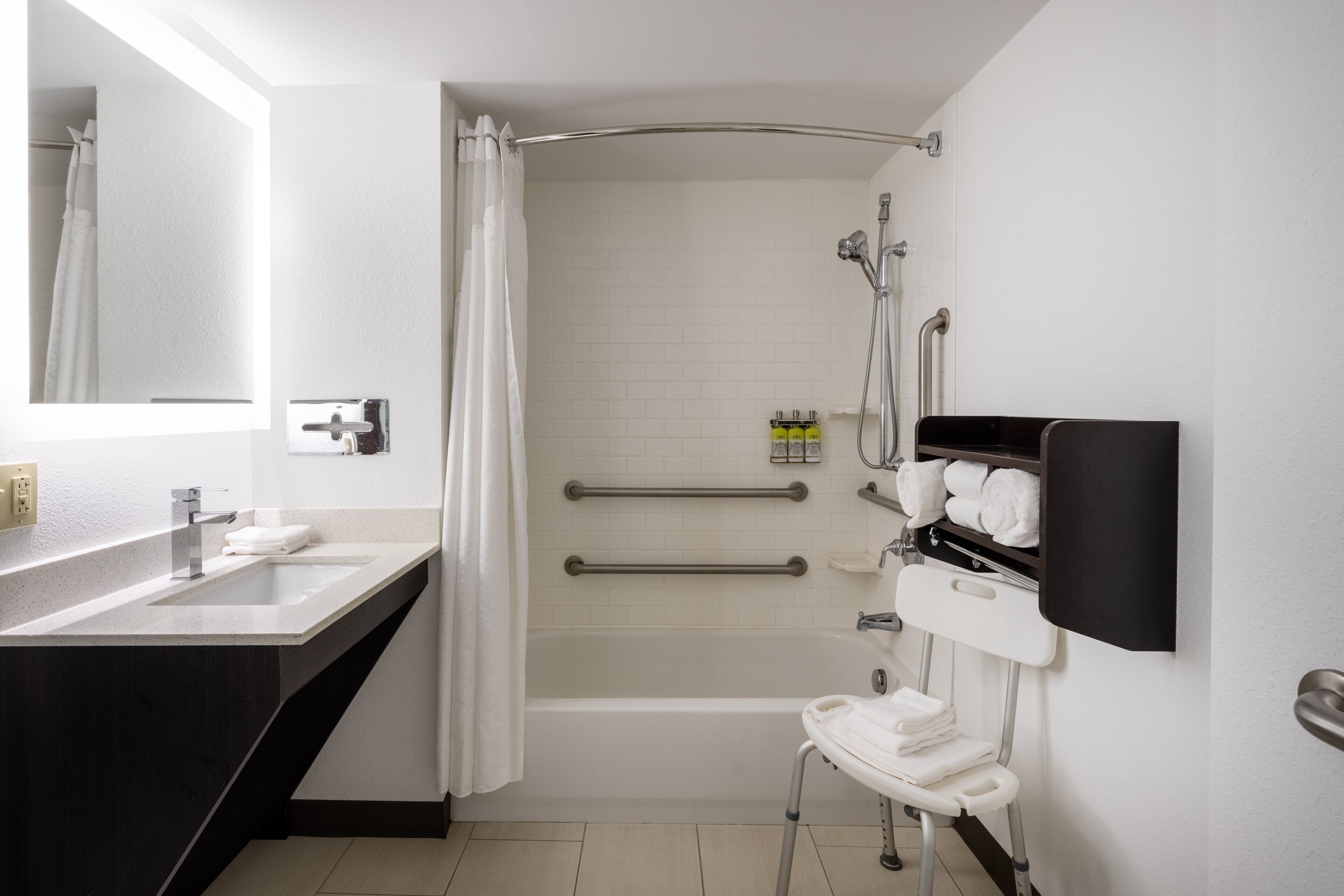 A modern bathroom with a bathtub, a shower, a toilet with a safety rail, a sink, and a mirror.