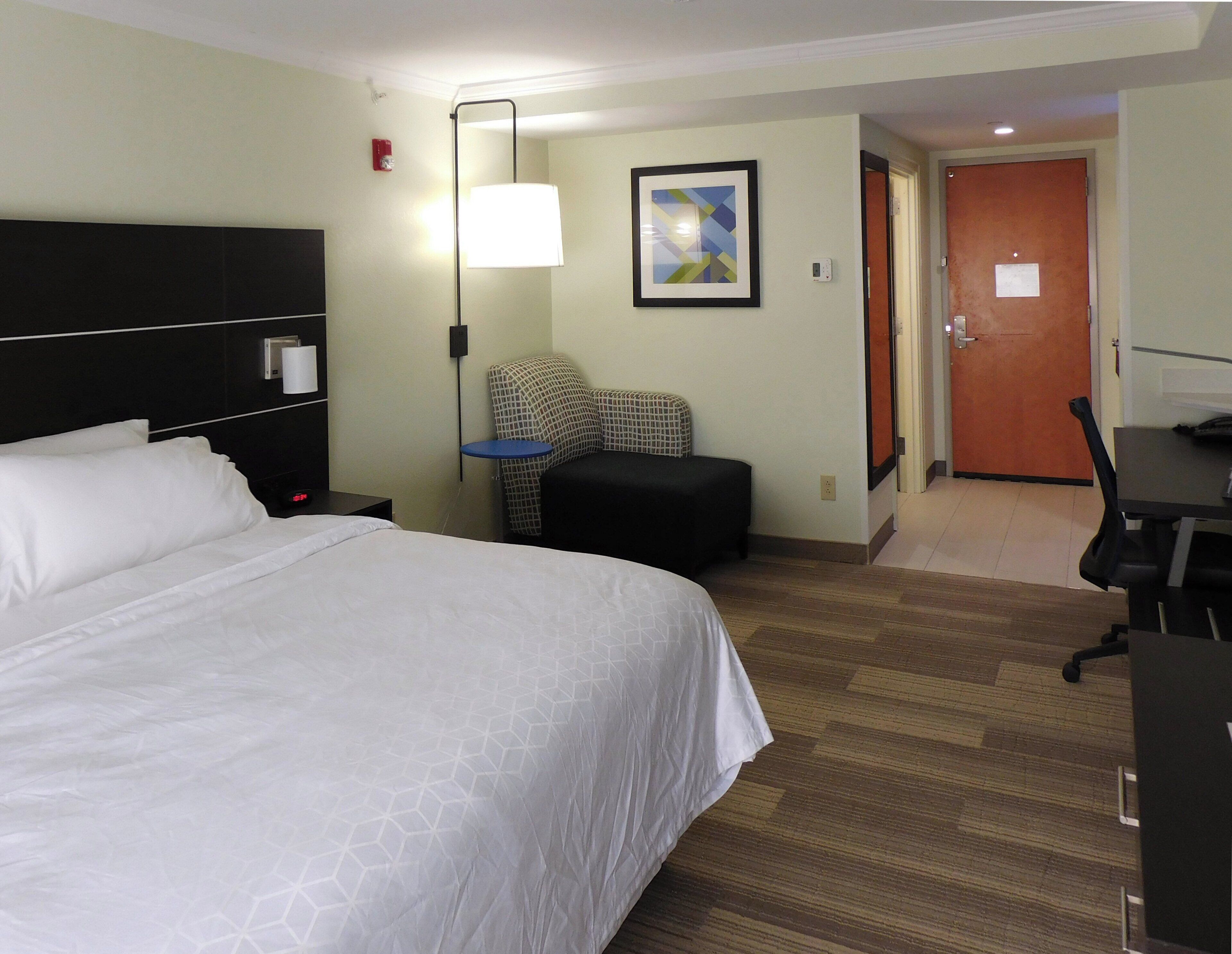 A hotel room with a large bed, a desk, a chair, a lamp, and a door.