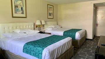 A hotel room with two beds, each with white linens and a green patterned throw. There are two wall-mounted lamps on a nightstand between the beds. The room has cream-colored walls and a door on the right.