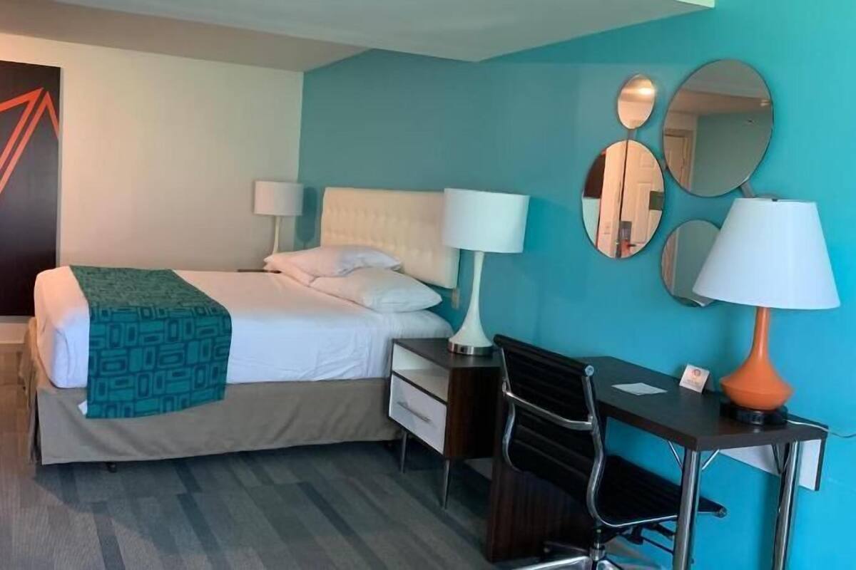 Habitación de hotel con cama, dos mesitas de noche con lámparas, un escritorio con silla y un cuadro abstracto grande en la pared.
