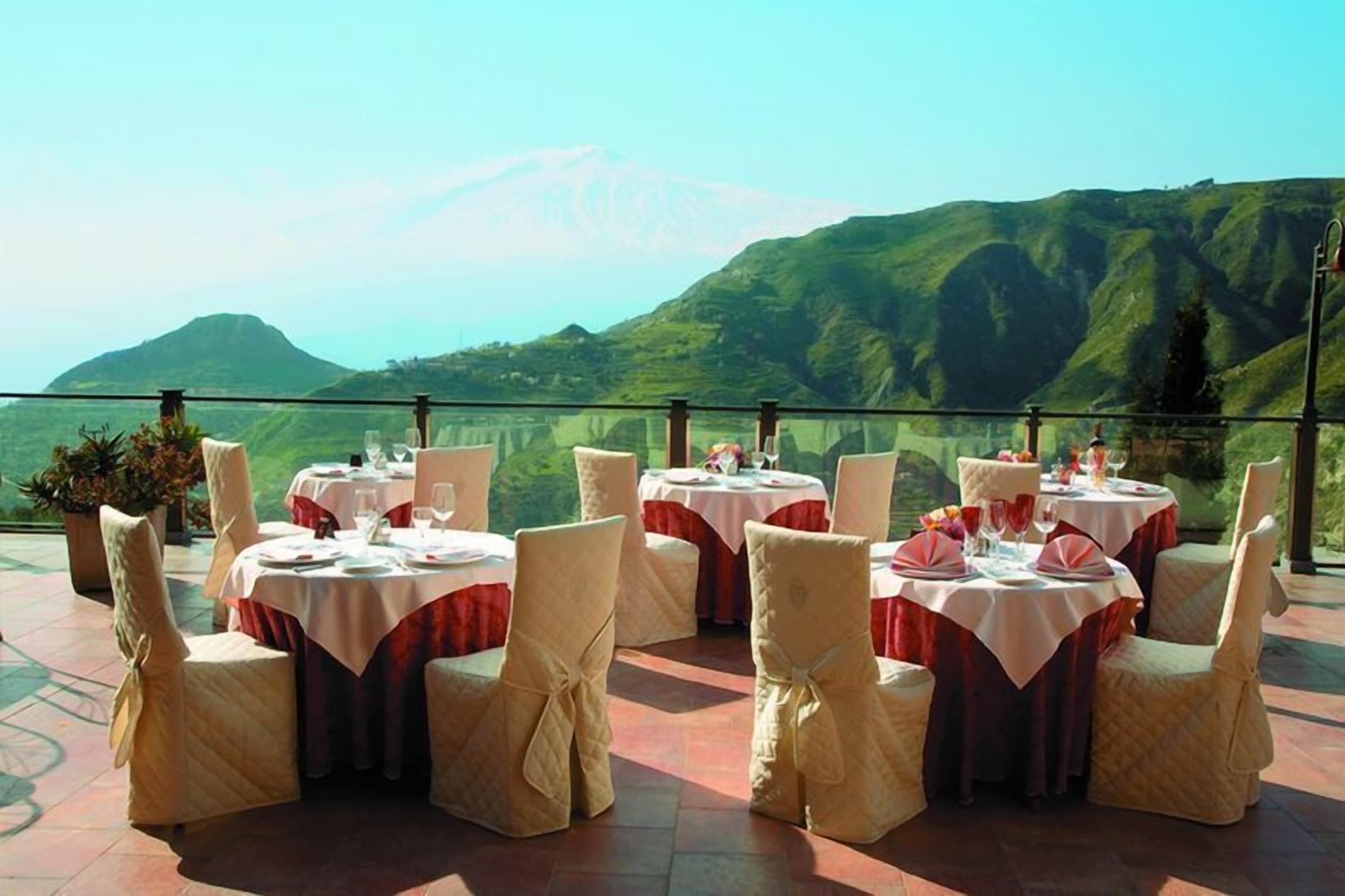 A dining area with tables set for six, each with white tablecloths and red accents, overlooking a mountainous landscape.