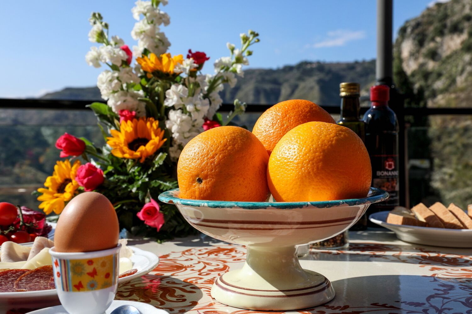A table with a bowl of oranges, a plate with bread, and a bottle of wine, set against a scenic mountain backdrop.