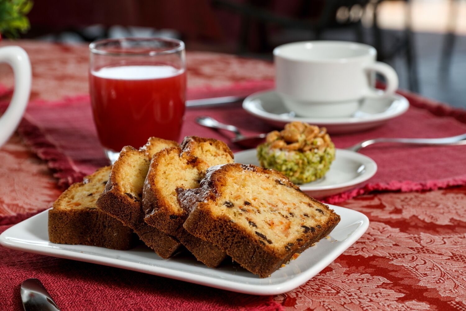 A slice of cake on a white plate, a glass of red beverage, and a small plate with a green dessert on a red tablecloth.