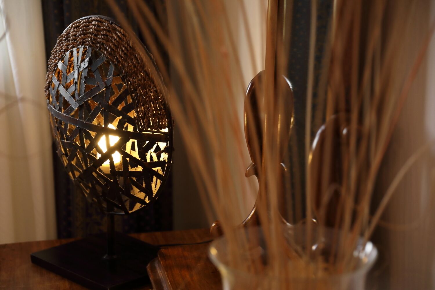 A decorative lamp with a spherical, mesh-like structure and a candle inside, placed on a wooden surface next to a vase with tall, slender objects.