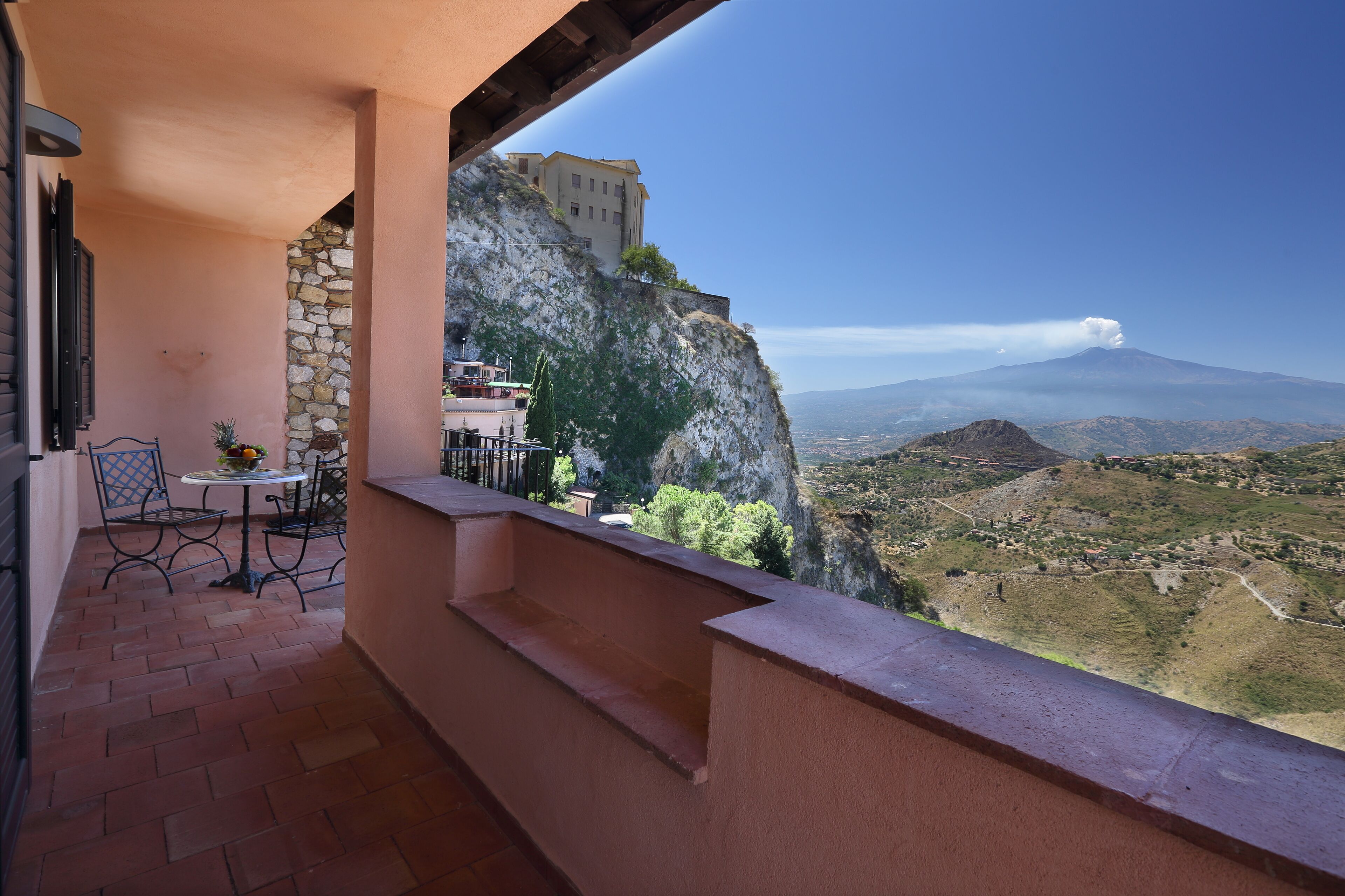 A balcony with a stone wall, a table with chairs, and a view of a mountain and a building.