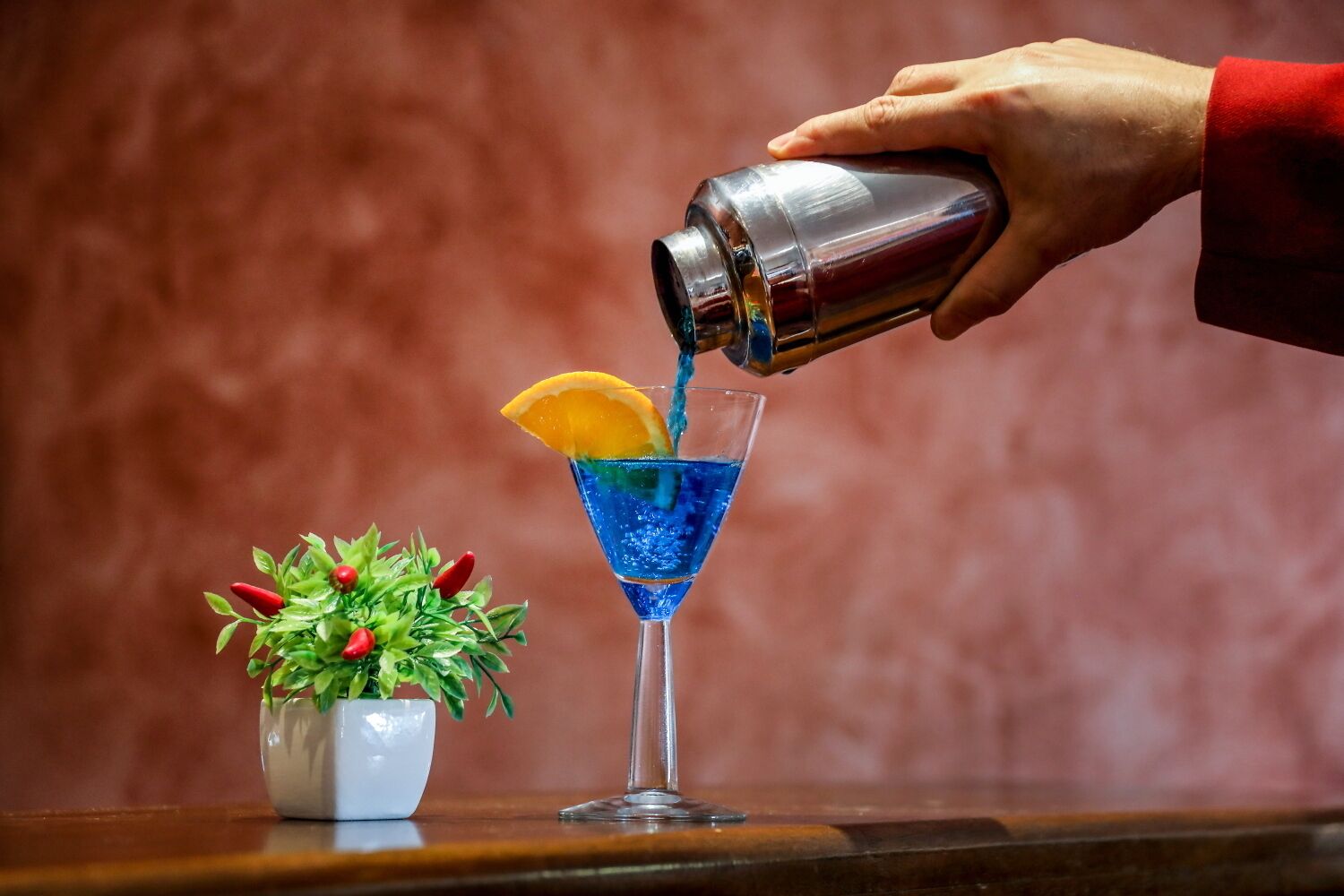 A hand pouring a blue liquid into a martini glass with an orange slice garnish.
