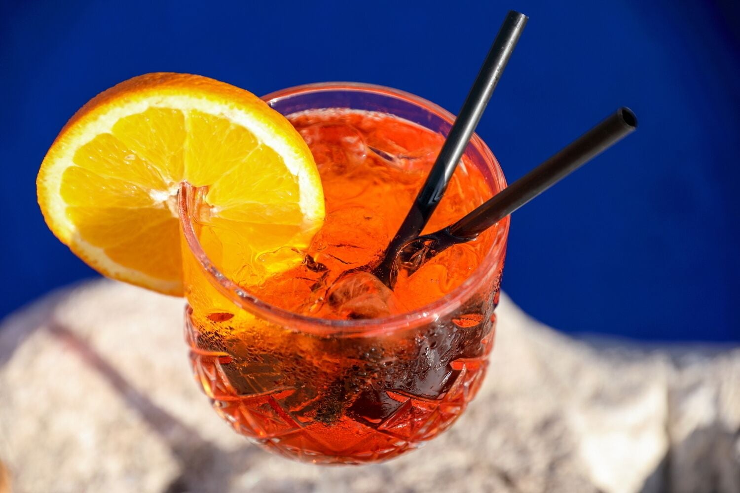 Un verre contenant une tranche d’orange et deux pailles, rempli d’une boisson rouge, le tout sur un fond bleu.