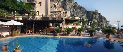 A pool area with lounge chairs and tables, surrounded by potted plants and a building with a balcony overlooking a cliff.