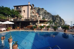 A pool area with lounge chairs and tables, surrounded by potted plants and a building with a balcony overlooking a cliff.