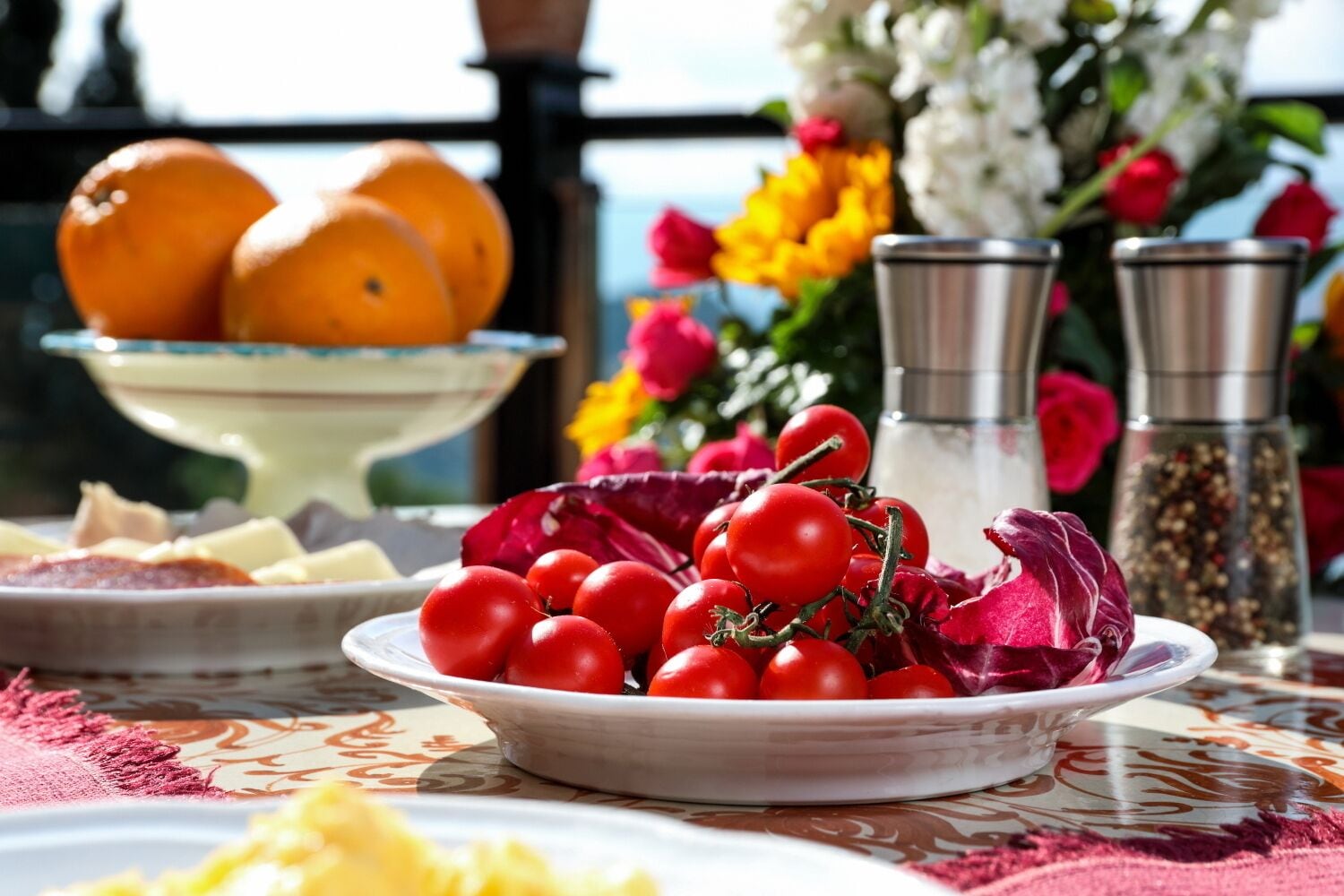 Una mesa con un plato de tomates cherry, un tazón de naranjas y un jarrón de flores.