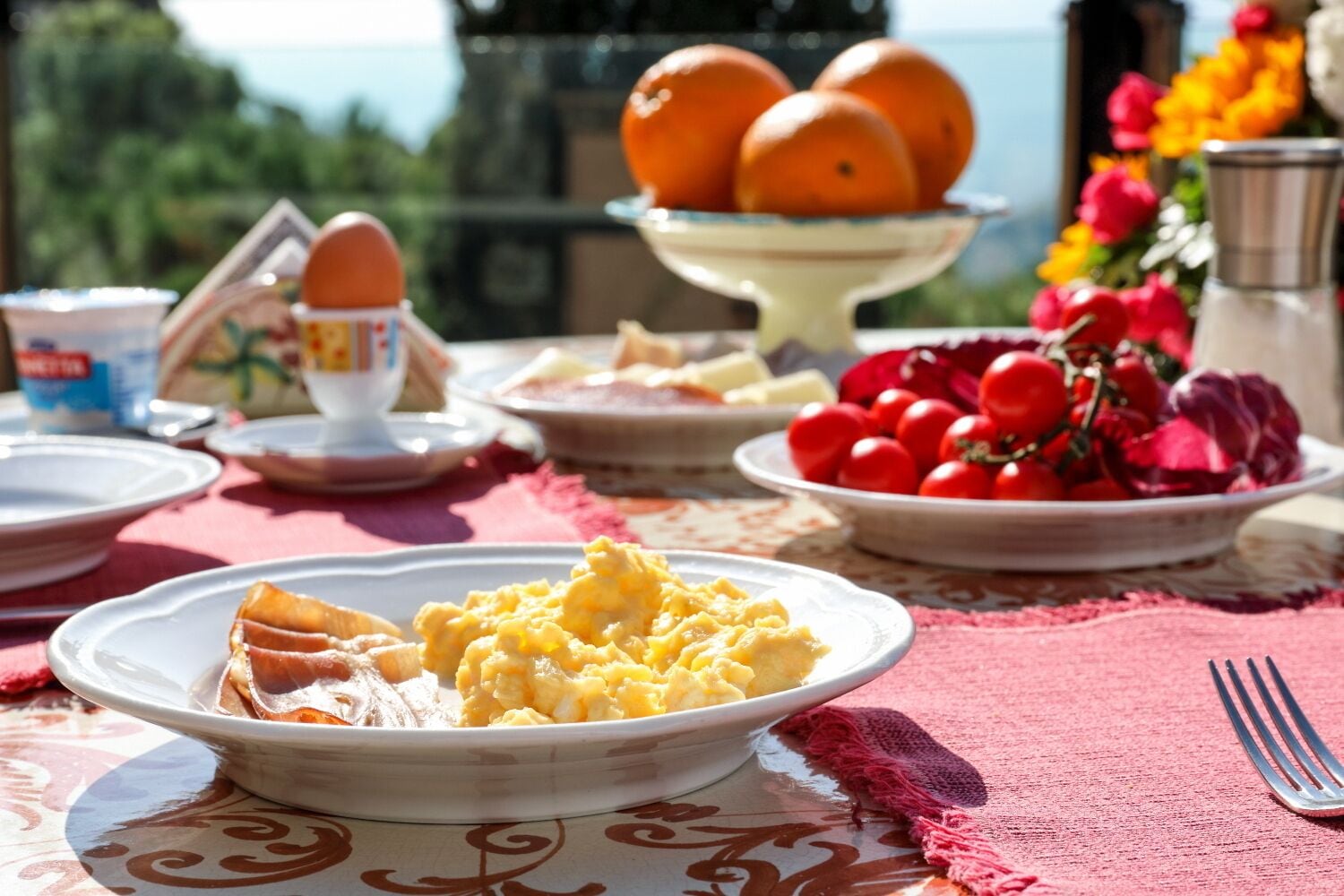 Una mesa de desayuno con platos de comida, un bol de naranjas y un florero con flores.