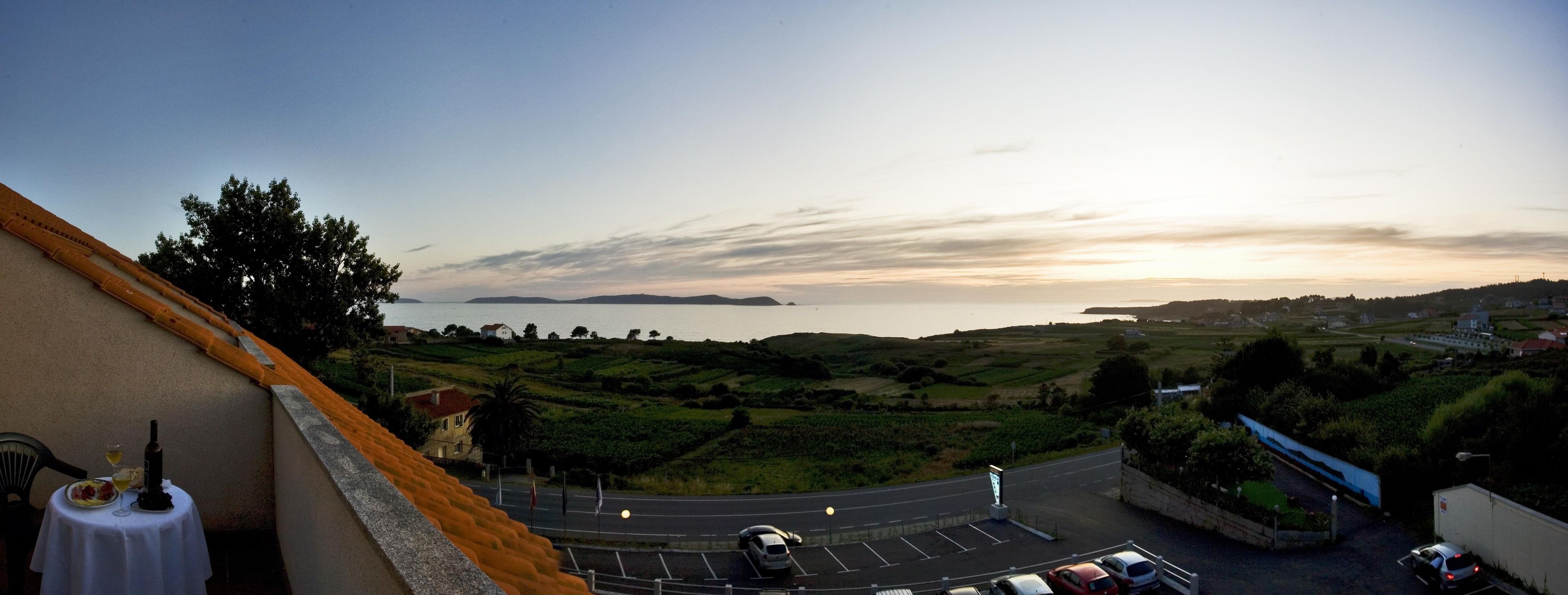 A coastal view with a road leading to a parking area, a building with a balcony overlooking the sea, and a distant island.