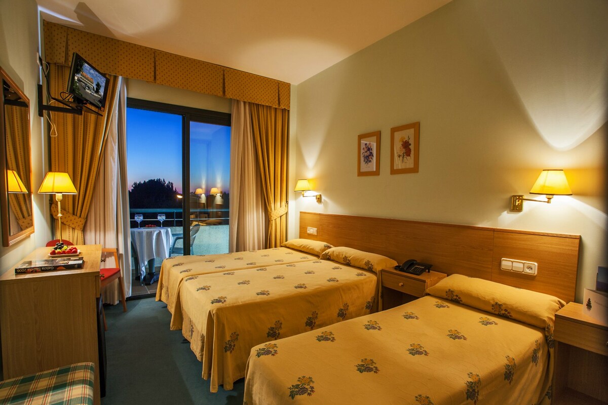 A hotel room with two beds, a desk with a lamp, a TV on the wall, and a view of the outdoors.