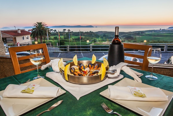 A table set for two with a seafood dish, wine glasses, and a bottle of wine, overlooking a scenic landscape.