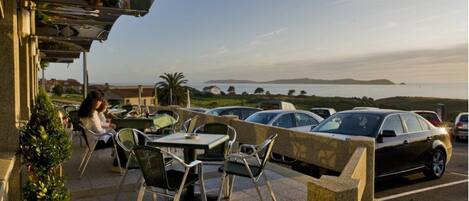 An outdoor cafe with tables and chairs, overlooking a parking area and a scenic view of the sea.