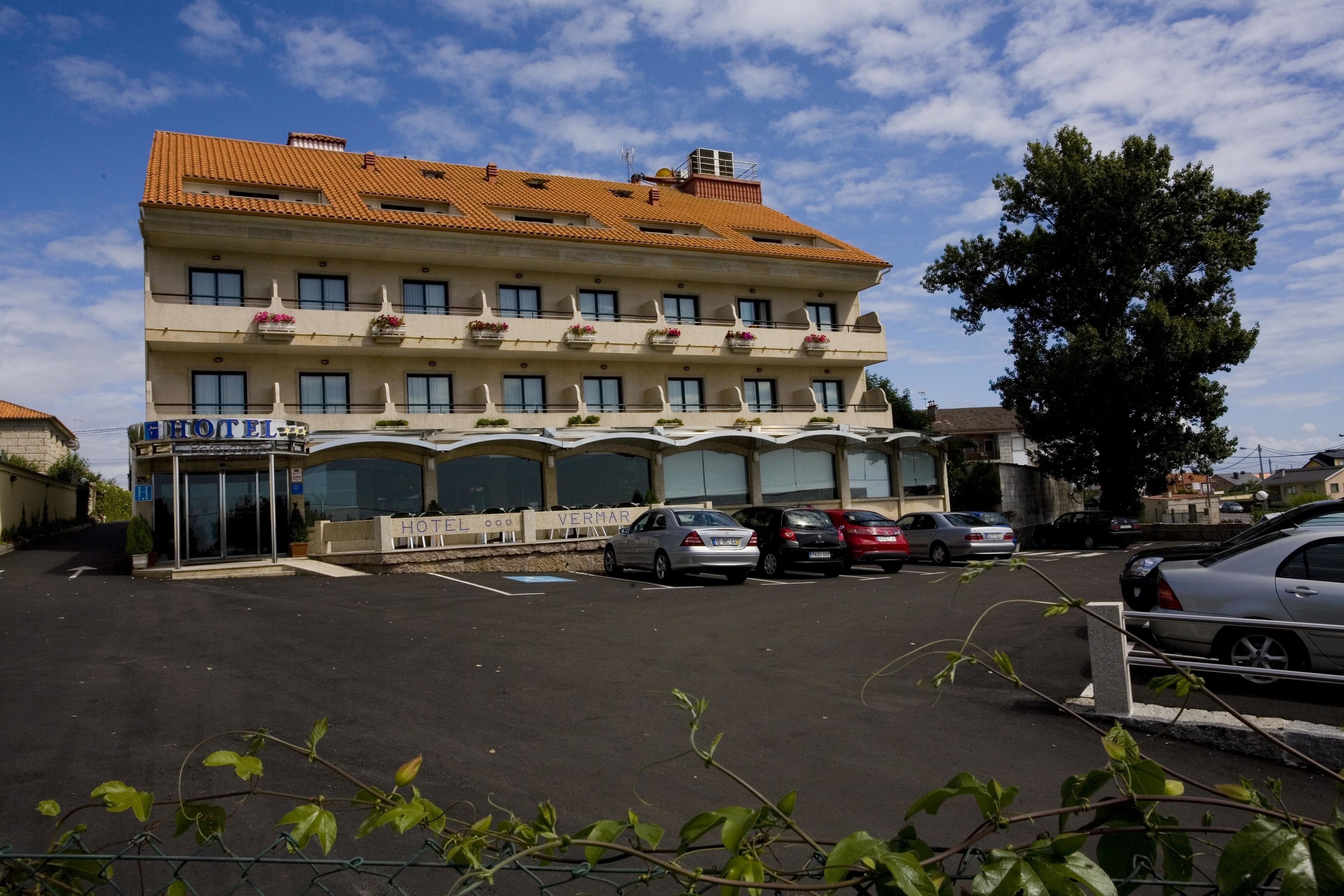 Un hotel con tejado de baldosas, múltiples balcones con jardineras y un estacionamiento con varios coches.
