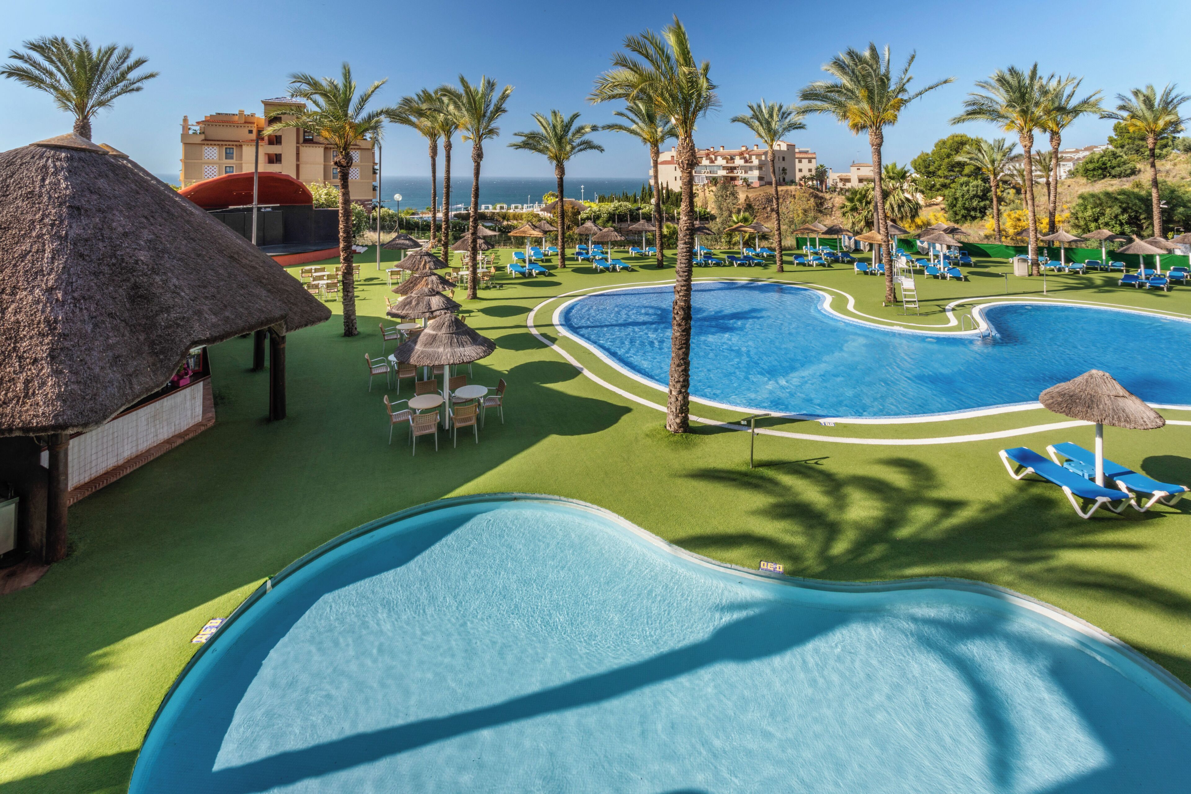 Un área de piscina en un resort con varias piscinas, sillas de descanso, palmeras y una estructura con techo de paja.
