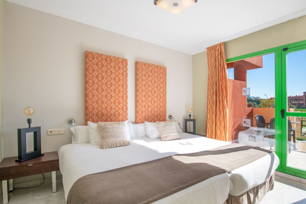 Una habitación de hotel con una cama grande, dos lámparas fijadas a la pared, una mesita de noche y vistas a través de una ventana con marco verde.