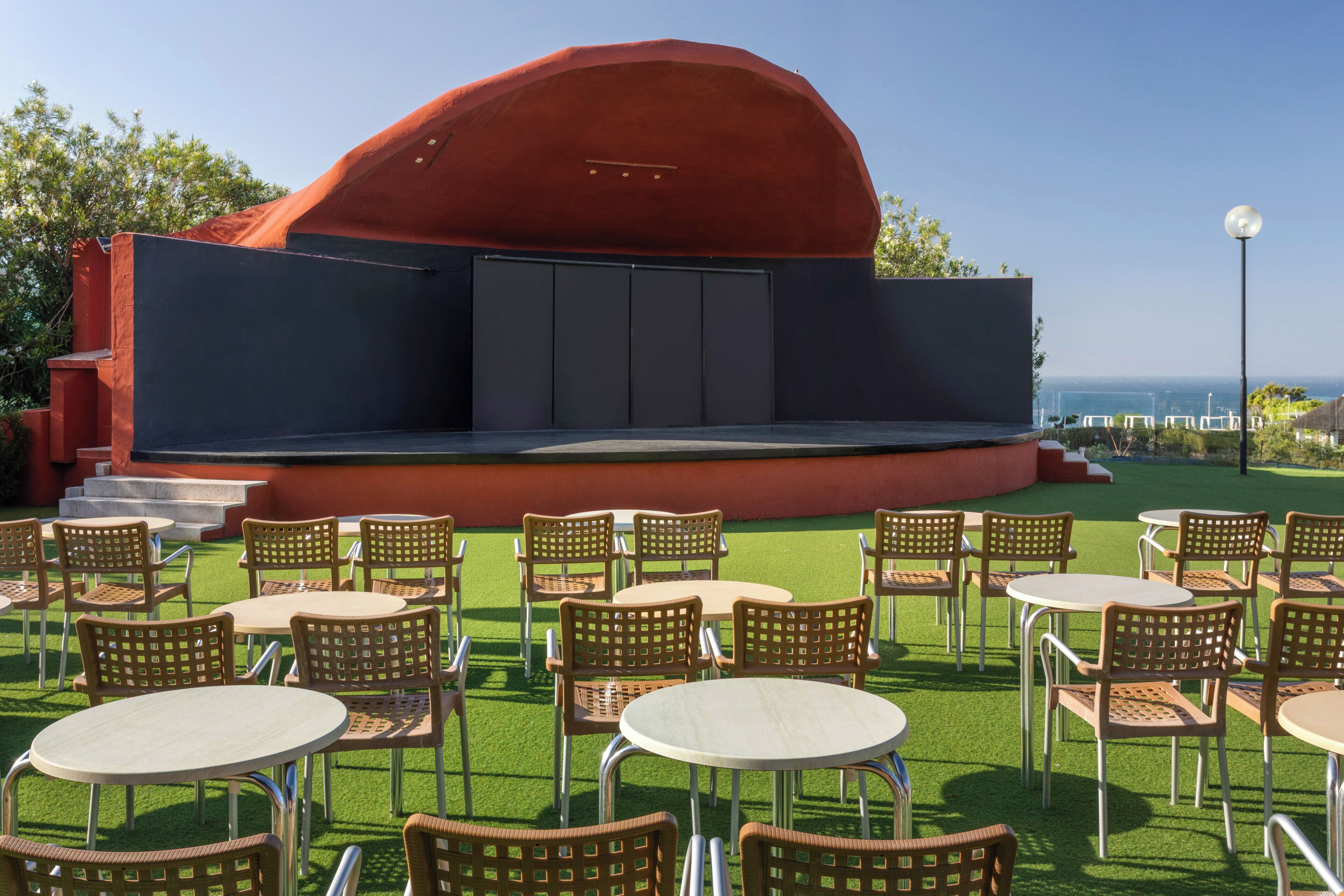 An outdoor seating area with tables and chairs, a stage, and a view of the sea.