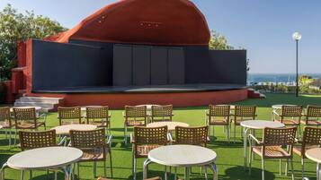 An outdoor seating area with tables and chairs, a stage, and a view of the sea.