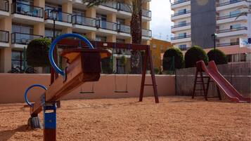 A playground with a swing set and a slide, surrounded by sand, with a building in the background.