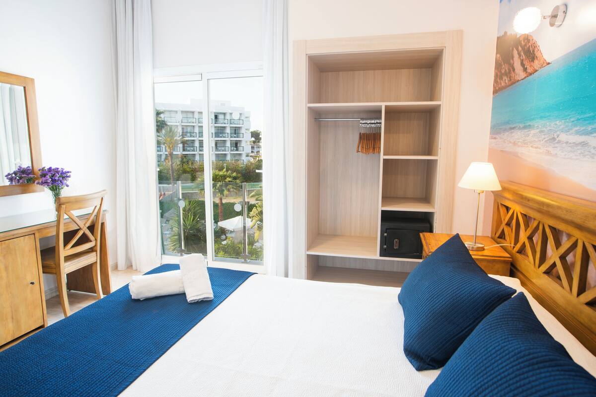 Habitación de hotel con una cama grande, un escritorio de madera, un armario y vistas a la ciudad desde un balcón.