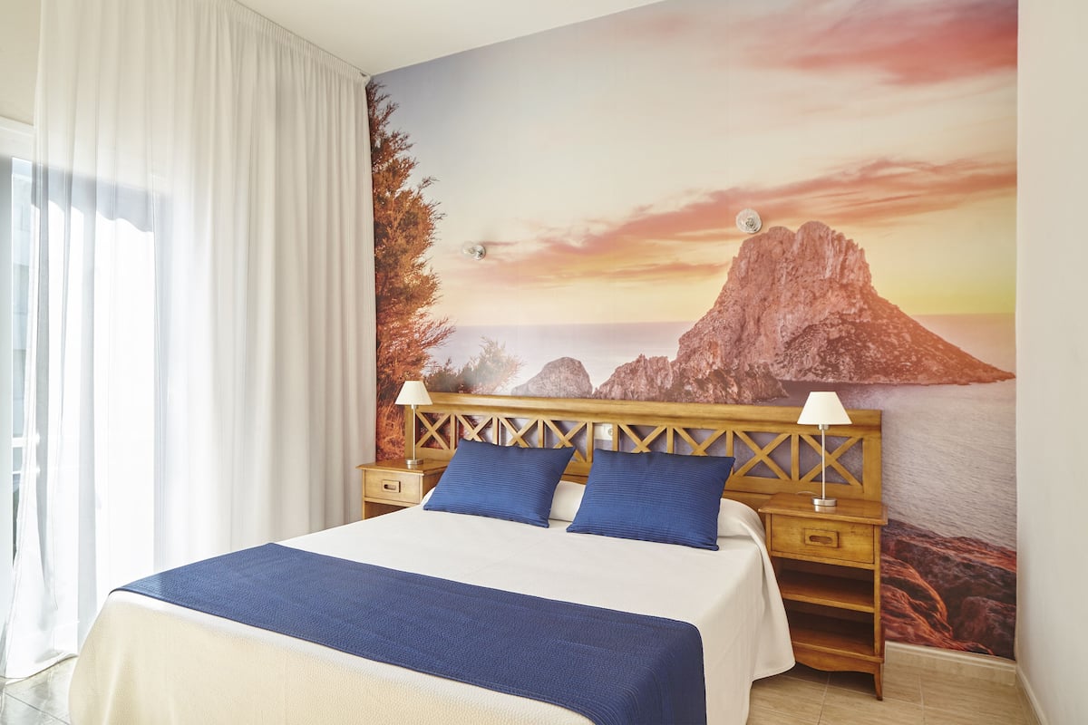 Una habitación de hotel con una cama, dos mesitas de noche, una lámpara y un mural grande en la pared que muestra una montaña y un atardecer.