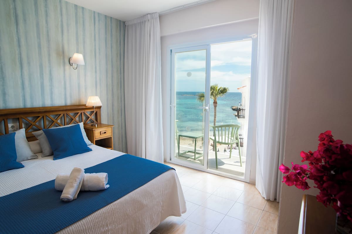 Habitación de hotel con una cama grande, una mesita de noche, una lámpara, vista al océano y un balcón con una silla y una mesa.