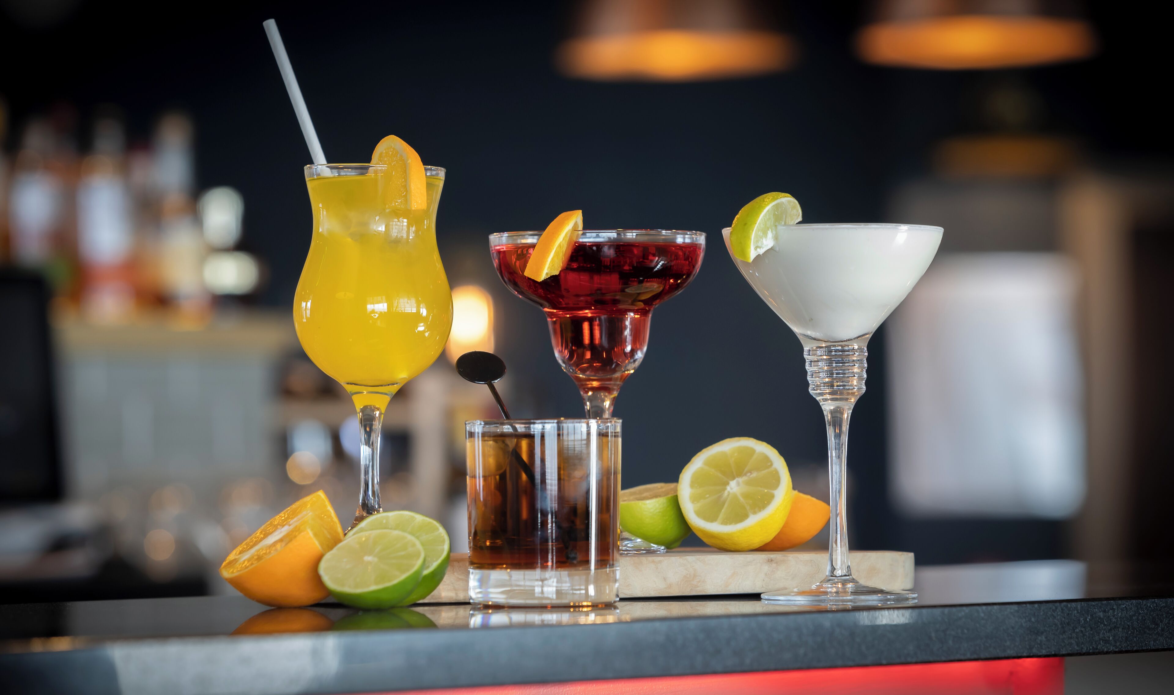 Three different cocktails on a bar counter with lime and orange slices beside them.