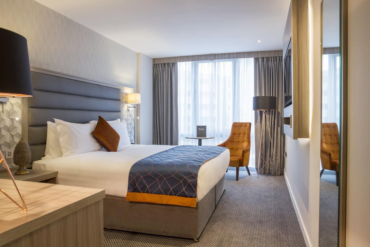 Habitación de hotel con una cama grande, un escritorio, dos sillas, una mesita pequeña y un espejo amplio.