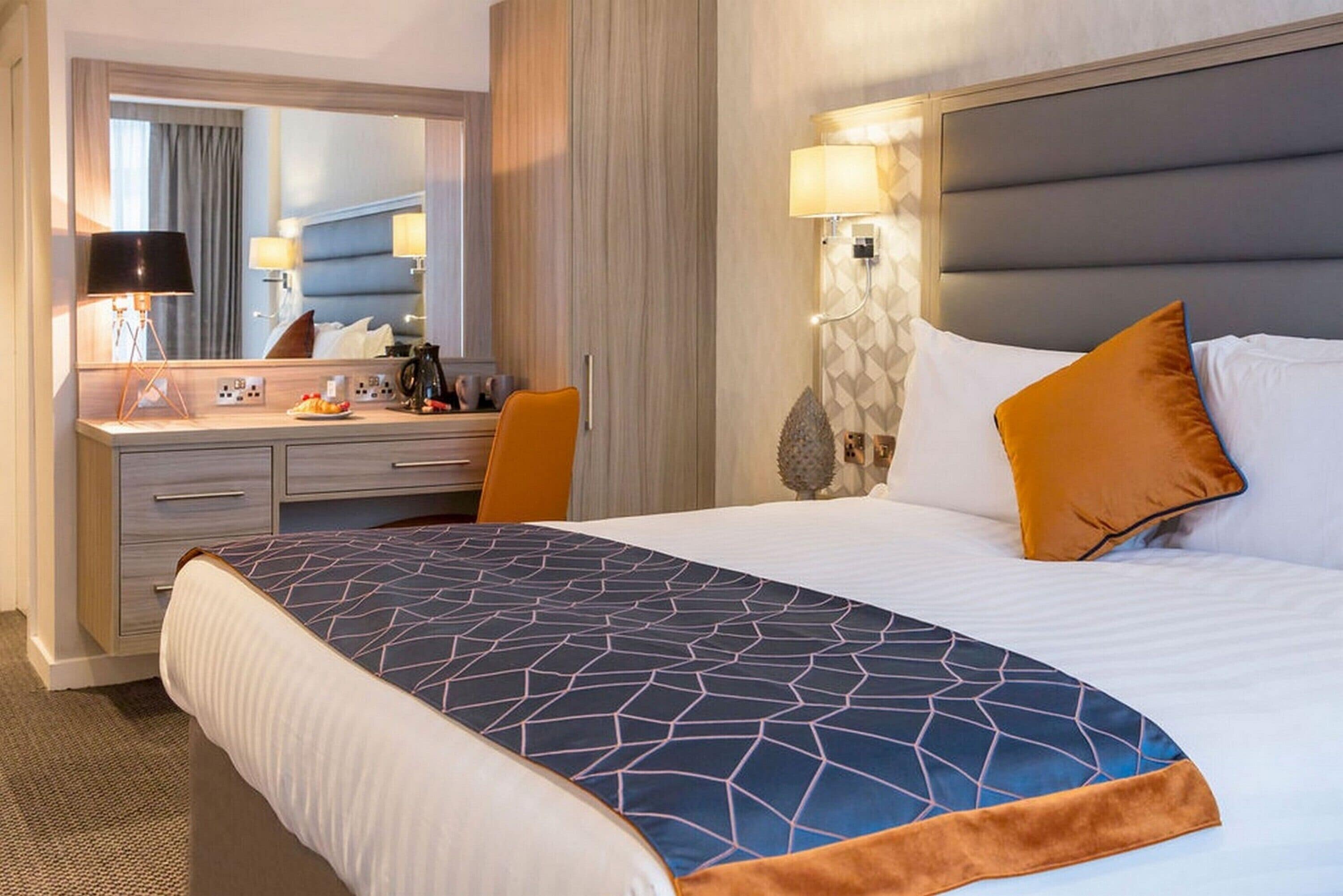 A hotel room with a large bed, a desk with a chair, a mirror, and a nightstand with a lamp.