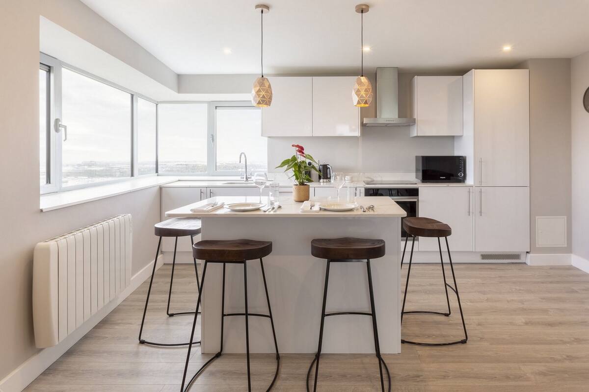 Una cocina moderna con una isla central, taburetes de bar, un microondas y vistas de la ciudad a través de amplios ventanales.
