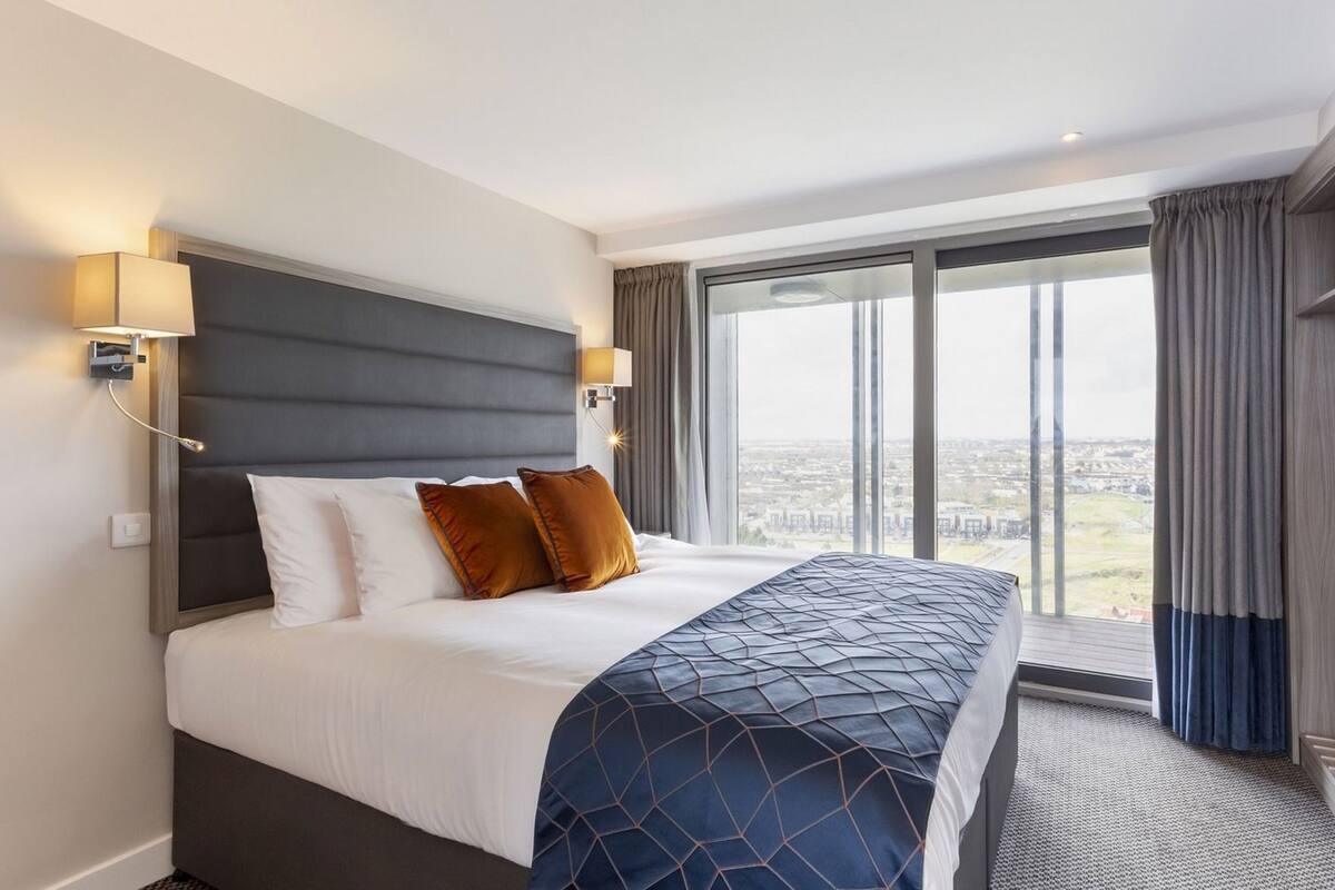 Habitación de hotel con una cama grande, dos lámparas de pared, vistas a la ciudad y un ventanal con cortinas.