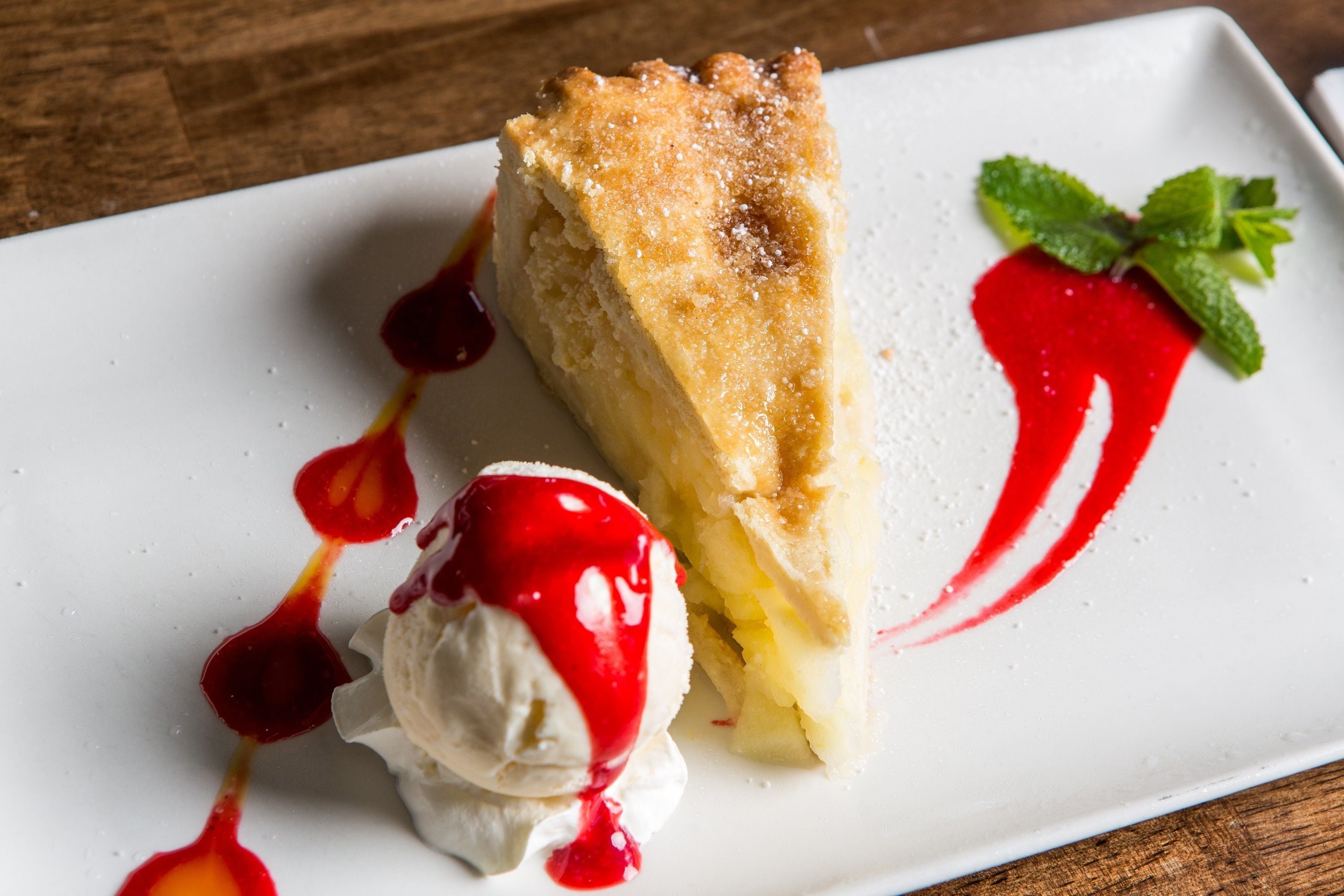 A slice of pie with a dusting of powdered sugar and a red sauce drizzle on a white plate.