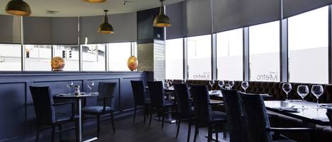 A restaurant with a modern interior, featuring dark wood furniture, pendant lighting, and large windows offering a view of the city.