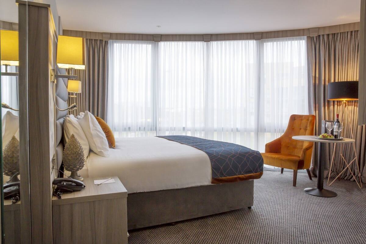 Habitación de hotel con una cama grande, un escritorio con teléfono, una silla, una mesita con una botella y vasos, y un ventanal con cortinas de encaje.