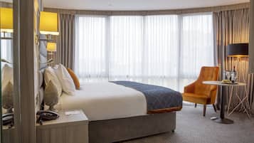 A hotel room with a large bed, a desk with a telephone, a chair, a small table with a bottle and glasses, and a large window with sheer curtains.