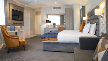 Habitación de hotel con una cama grande, un sofá, un escritorio con silla, un televisor montado en la pared y una ventana con cortinas.