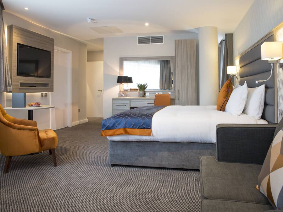 Habitación de hotel con una cama grande, un sofá, un escritorio con silla, un televisor montado en la pared y una ventana con cortinas.