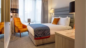 Habitación de hotel con cama, escritorio, silla, lámpara y vistas al exterior.